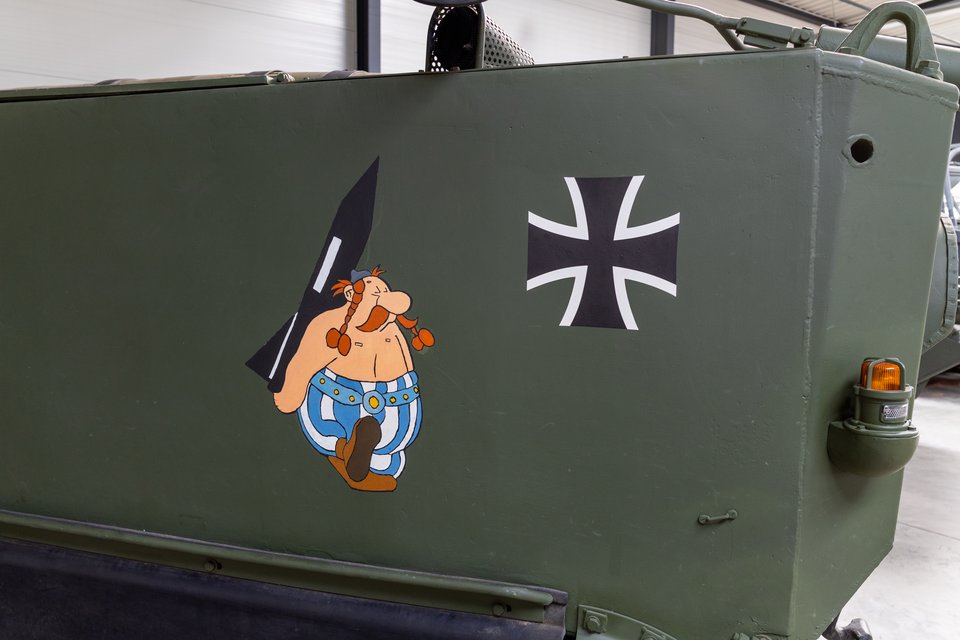 Side panel of an olive-green military armored vehicle featuring a painted cartoon figure with red braided mustache holding a menhir, next to a black and white Iron Cross emblem and an orange marker light.