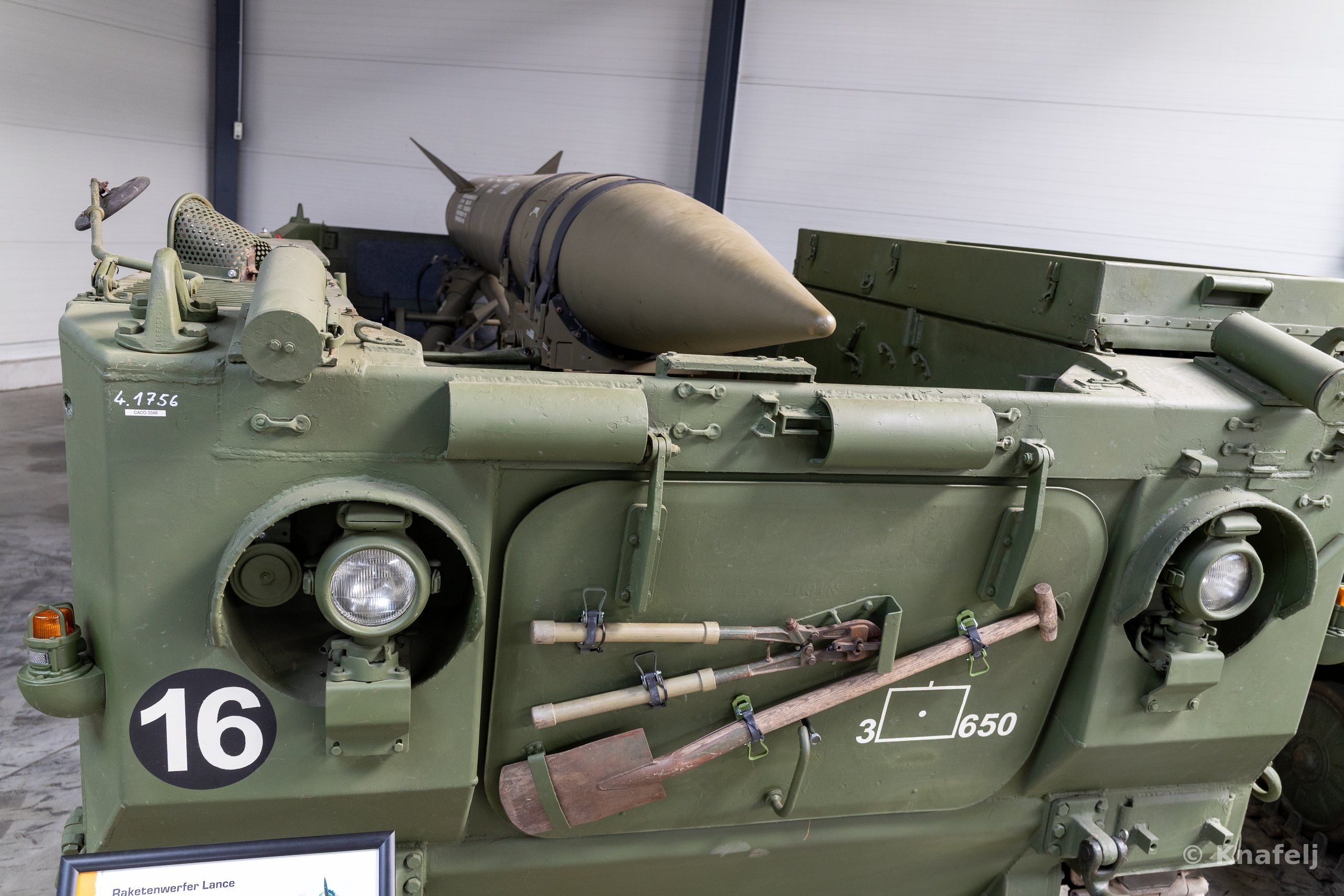 Front section of a green tracked military vehicle bearing a white number 16 on a black circle, with a missile mounted on top, entrenching tools and equipment strapped to the front panel, and round headlights, displayed indoors.