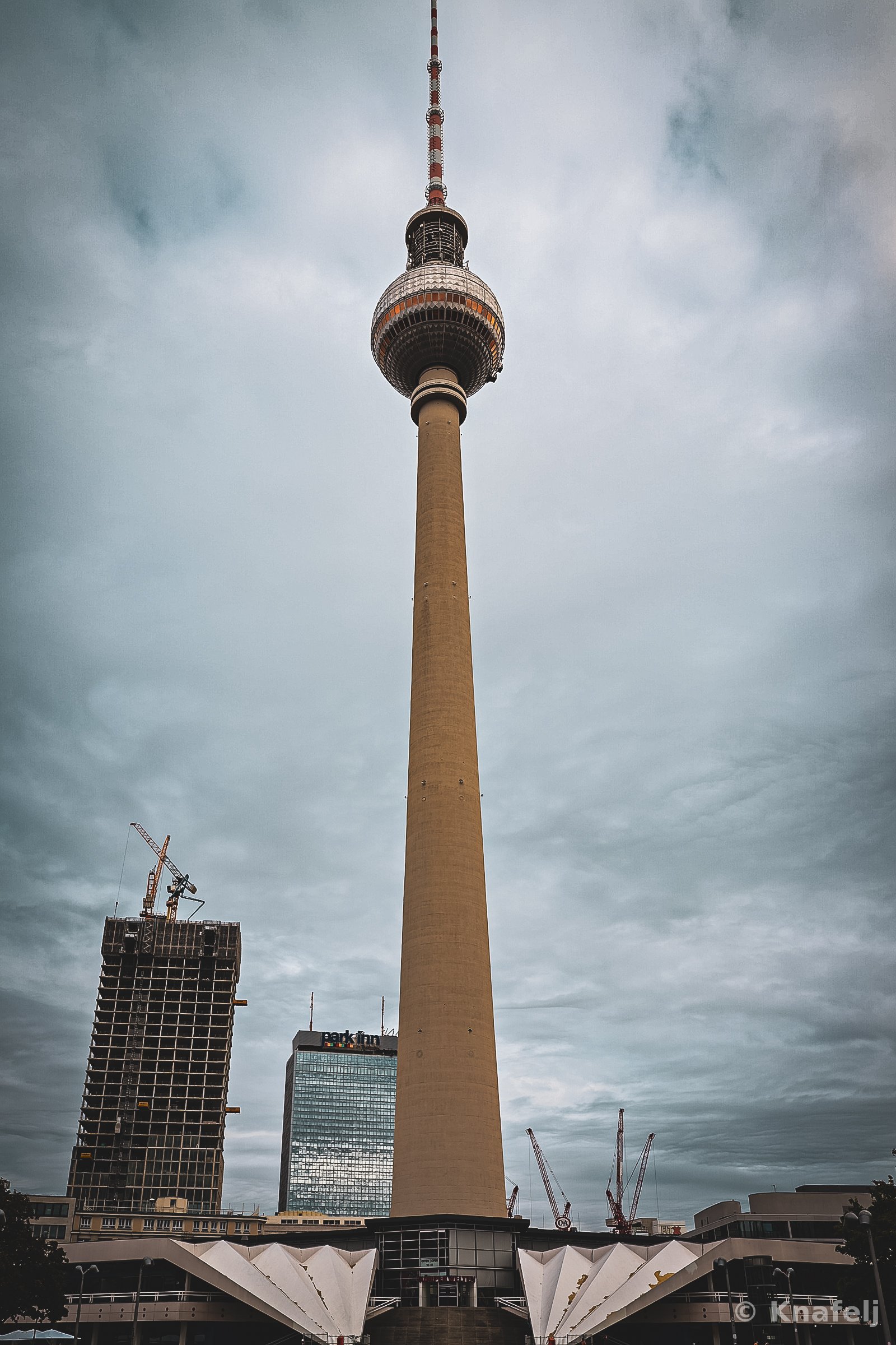 A tall television tower with a spherical observation deck stands against a cloudy grey sky, surrounded by glass office buildings and construction cranes in the city below.