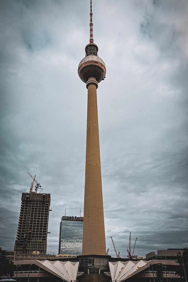 A tall television tower with a spherical observation deck stands against a cloudy grey sky, surrounded by glass office buildings and construction cranes in the city below.