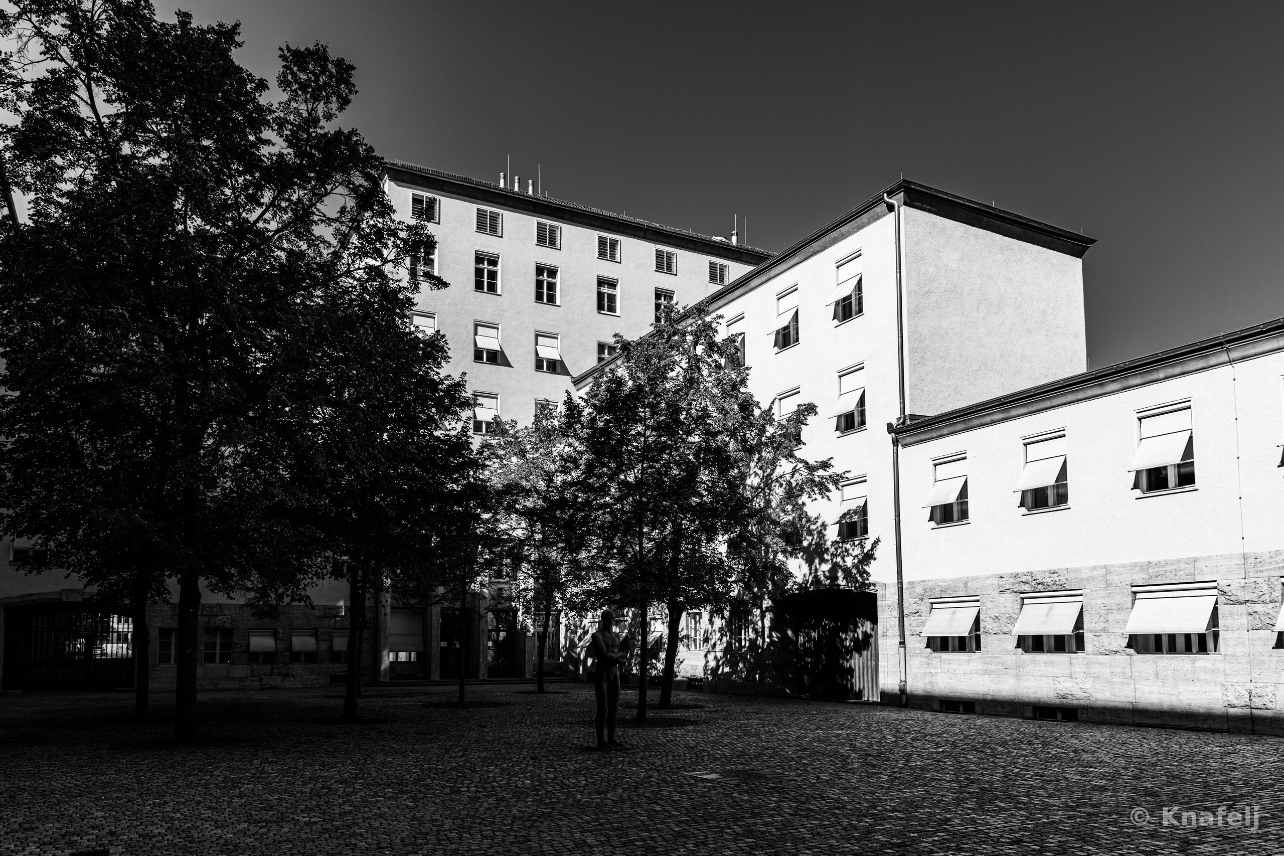 Black and white photo of a person standing alone in a cobblestone courtyard, flanked by trees and surrounded by large multi-story buildings with rows of windows under a clear sky.