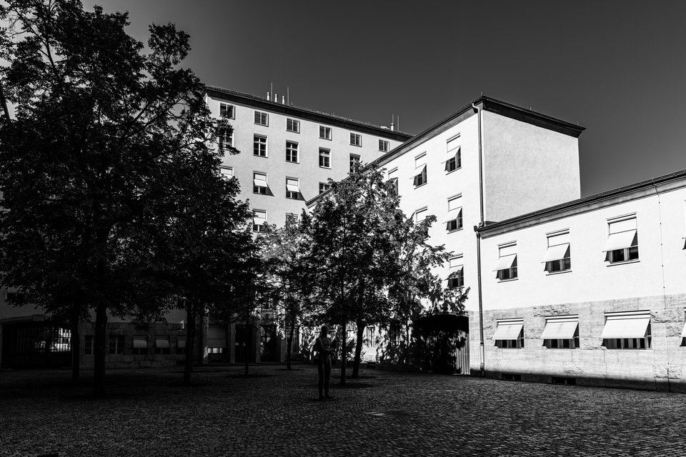 Black and white photo of a person standing alone in a cobblestone courtyard, flanked by trees and surrounded by large multi-story buildings with rows of windows under a clear sky.