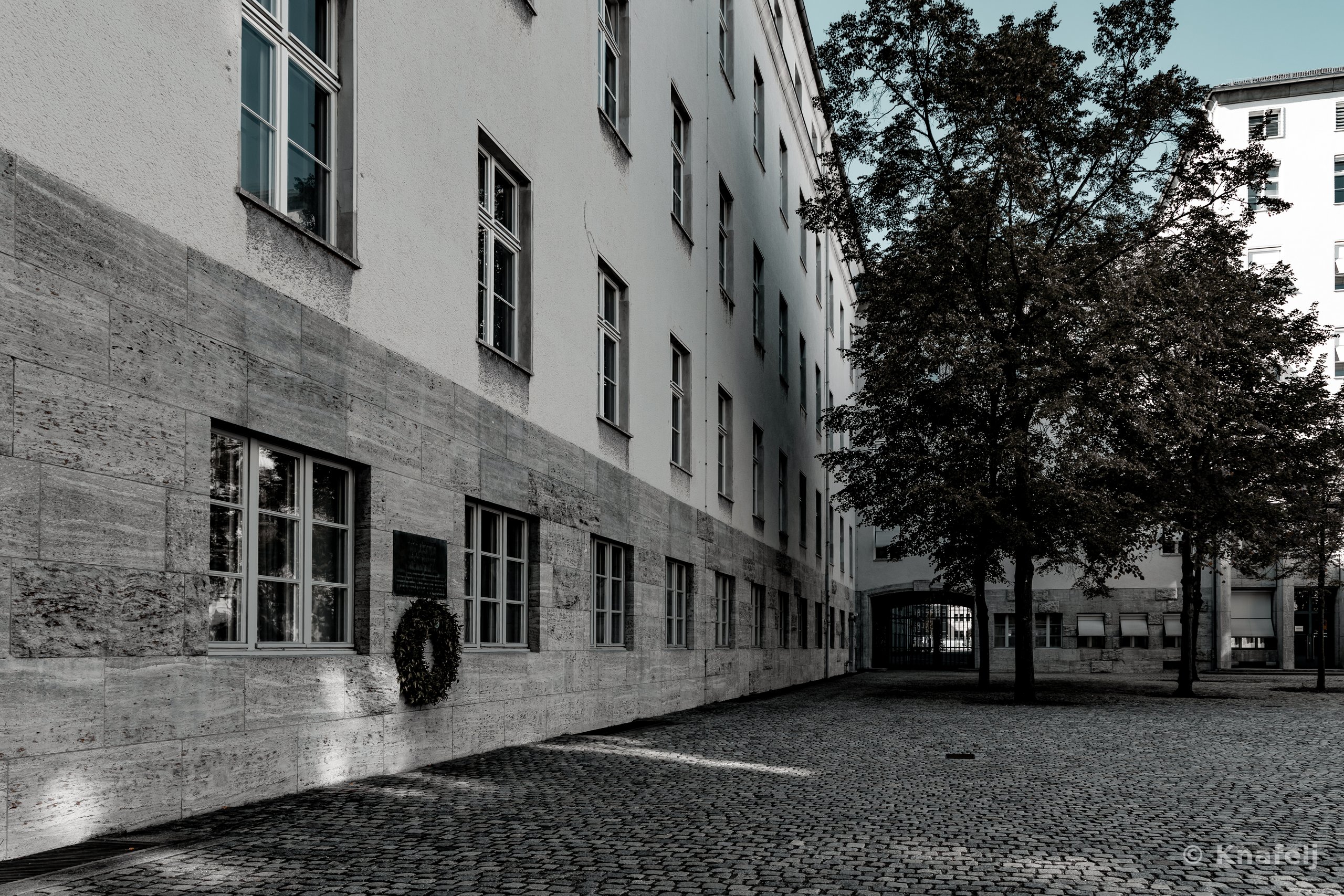A cobblestone courtyard beside a tall building with a stone facade and many windows. A dark memorial plaque with a wreath is mounted on the lower wall, and trees stand on the right against another building.