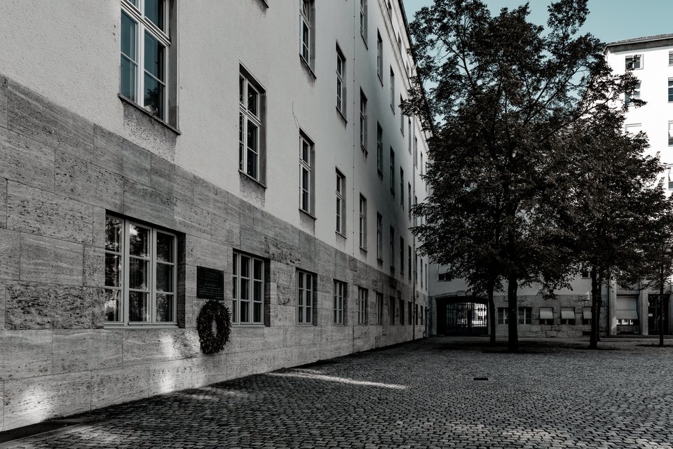 A cobblestone courtyard beside a tall building with a stone facade and many windows. A dark memorial plaque with a wreath is mounted on the lower wall, and trees stand on the right against another building.