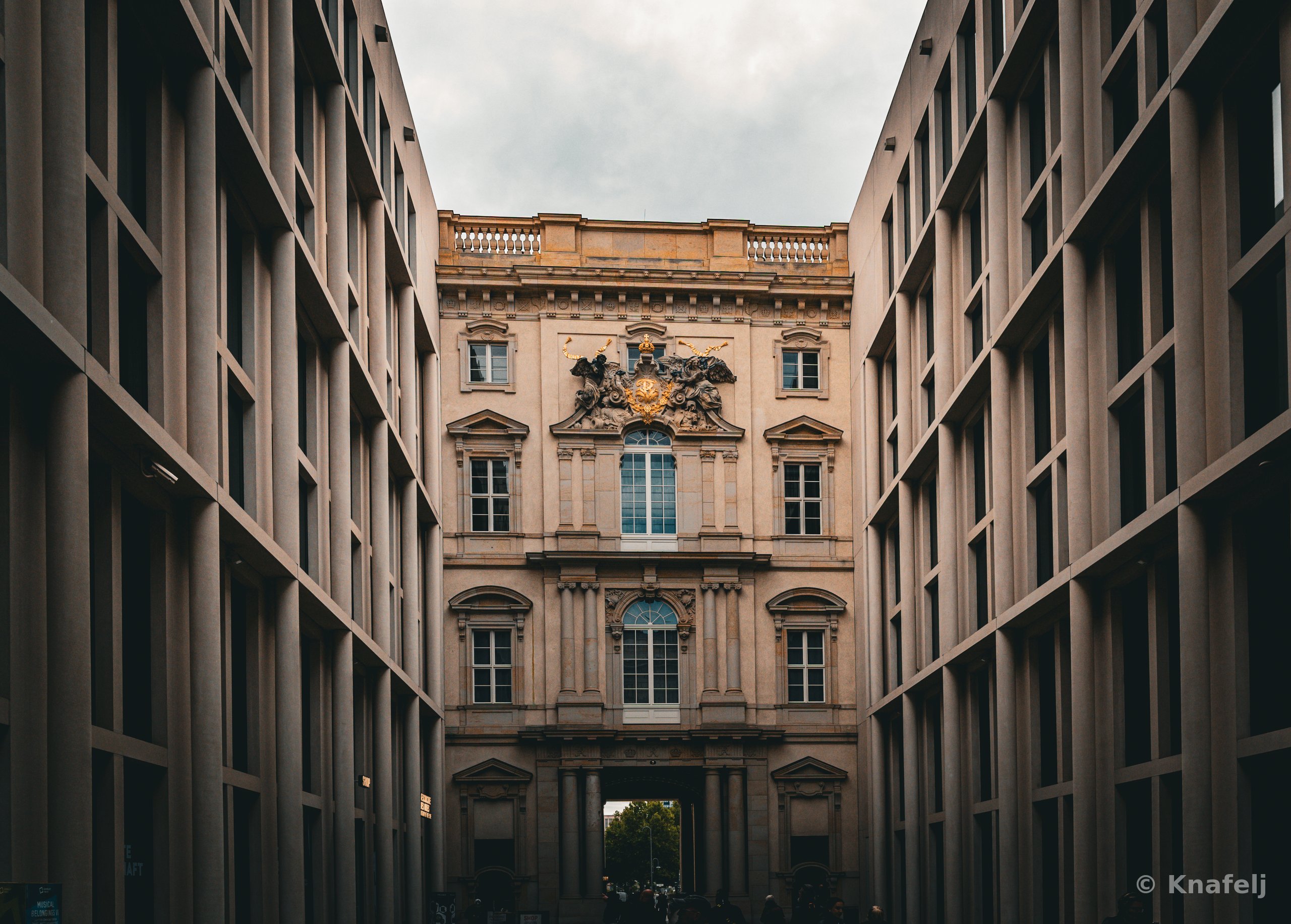 A historic baroque building with columns, arched windows, and golden ornamentation seen at the end of a corridor formed by two modern gray-windowed buildings on either side, beneath a cloudy sky.