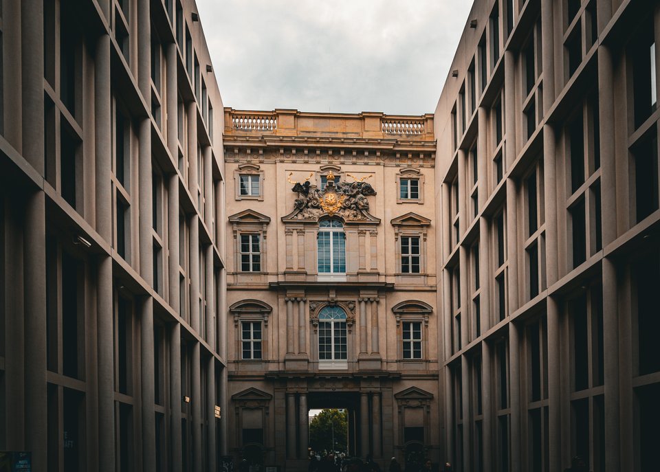 A historic baroque building with columns, arched windows, and golden ornamentation seen at the end of a corridor formed by two modern gray-windowed buildings on either side, beneath a cloudy sky.