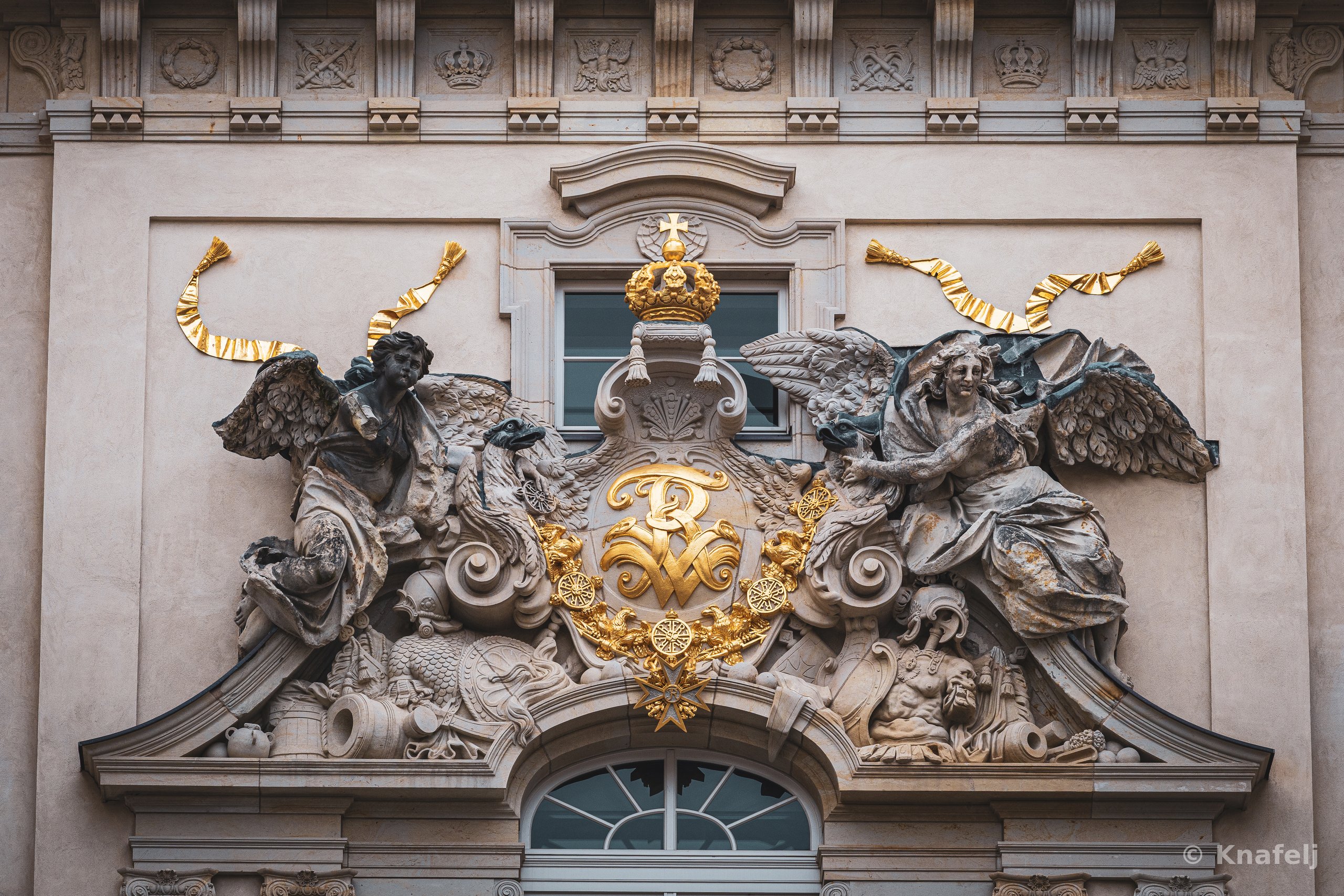 Detailed Baroque architectural relief on a building facade showing two winged angel statues on either side of a golden coat of arms with a crown and ornate decorations above an arched window.