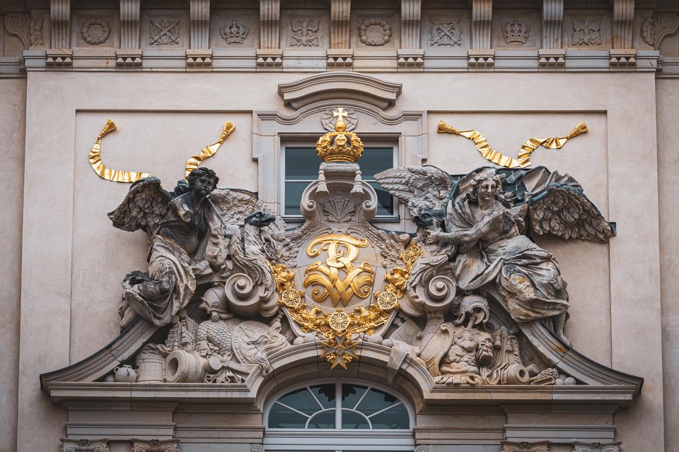 Detailed Baroque architectural relief on a building facade showing two winged angel statues on either side of a golden coat of arms with a crown and ornate decorations above an arched window.