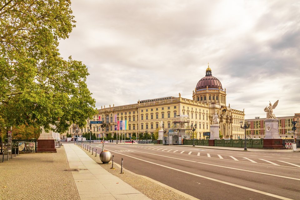 A wide street leading toward a large yellow neoclassical palace with a domed tower and a cross on top, with statues on bridge pillars to the right and a tree on the left under an overcast sky.