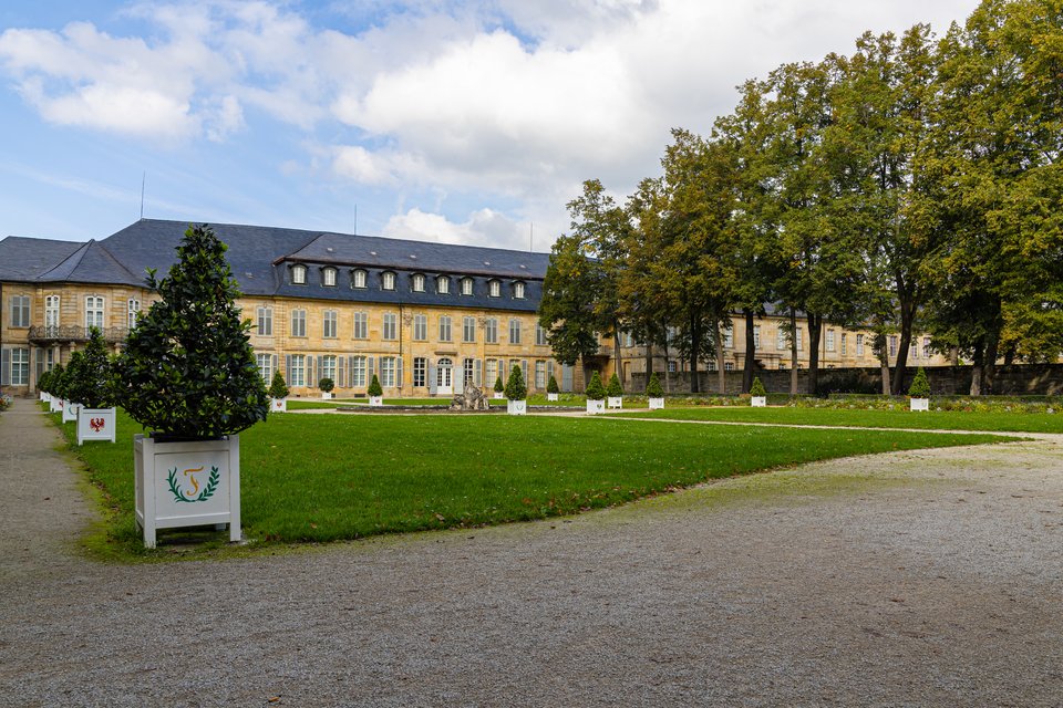 A large yellow baroque palace with slate roofs sits behind a green lawn and gravel courtyard, lined with white planter boxes holding cone-shaped topiary trees, surrounded by tall trees under a cloudy blue sky.