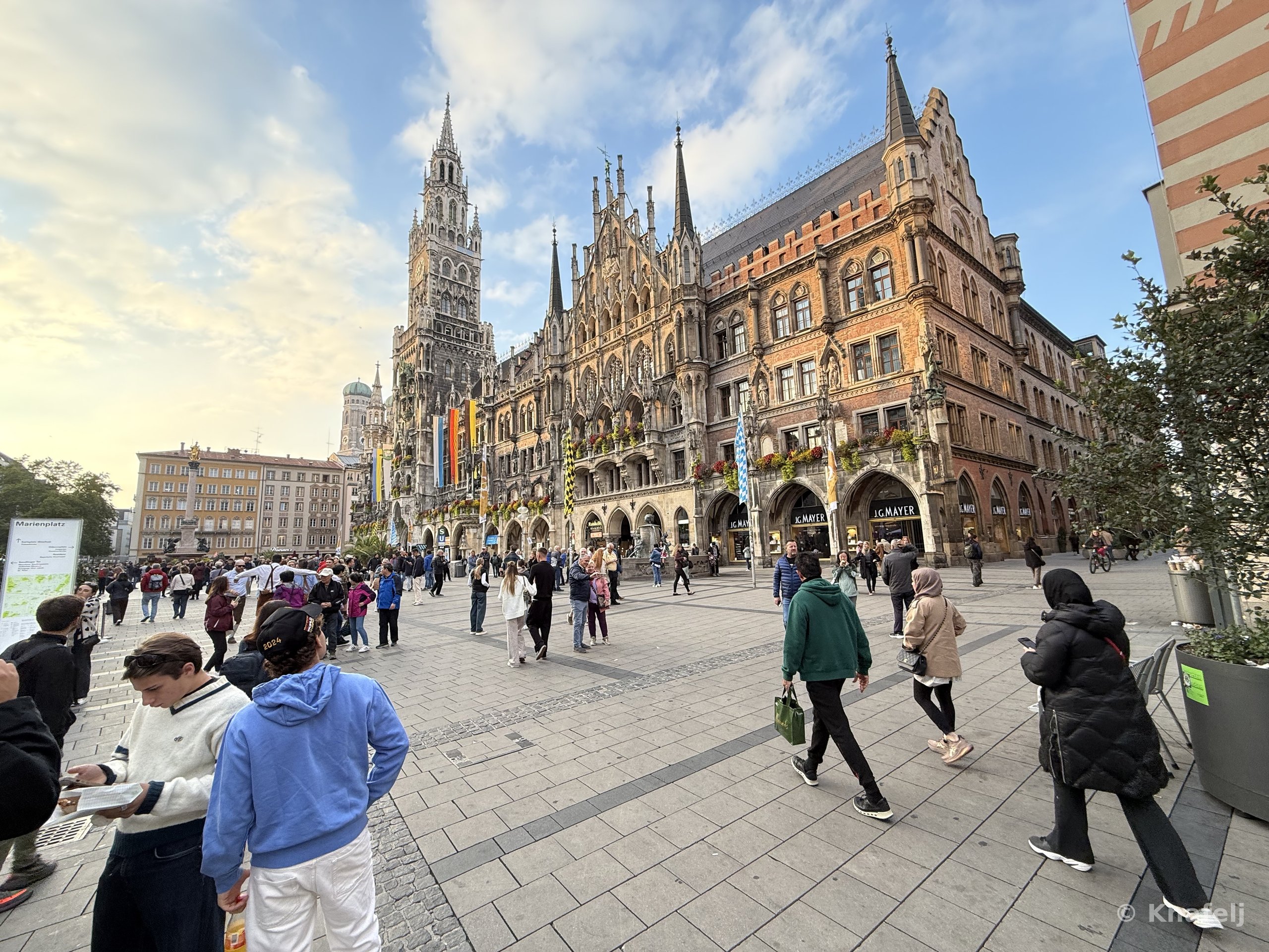 A crowded city square with people walking on cobblestone pavement in front of a large Gothic-style town hall with a tall clock tower under a partly cloudy sky.