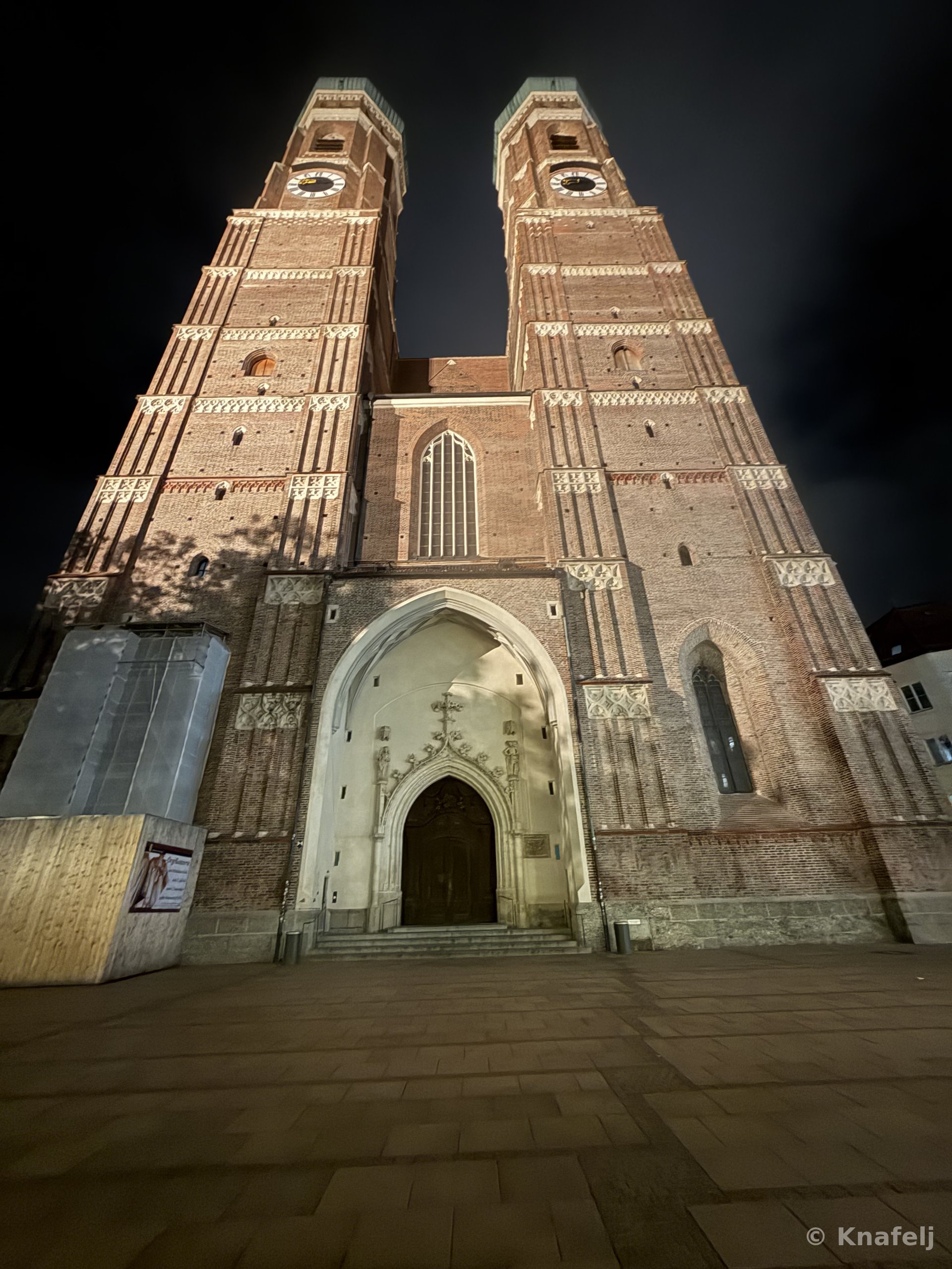 Low-angle night view of a tall Gothic brick cathedral with two towers and clock faces, lit warmly against a dark sky, with stone steps leading to an arched wooden doorway.