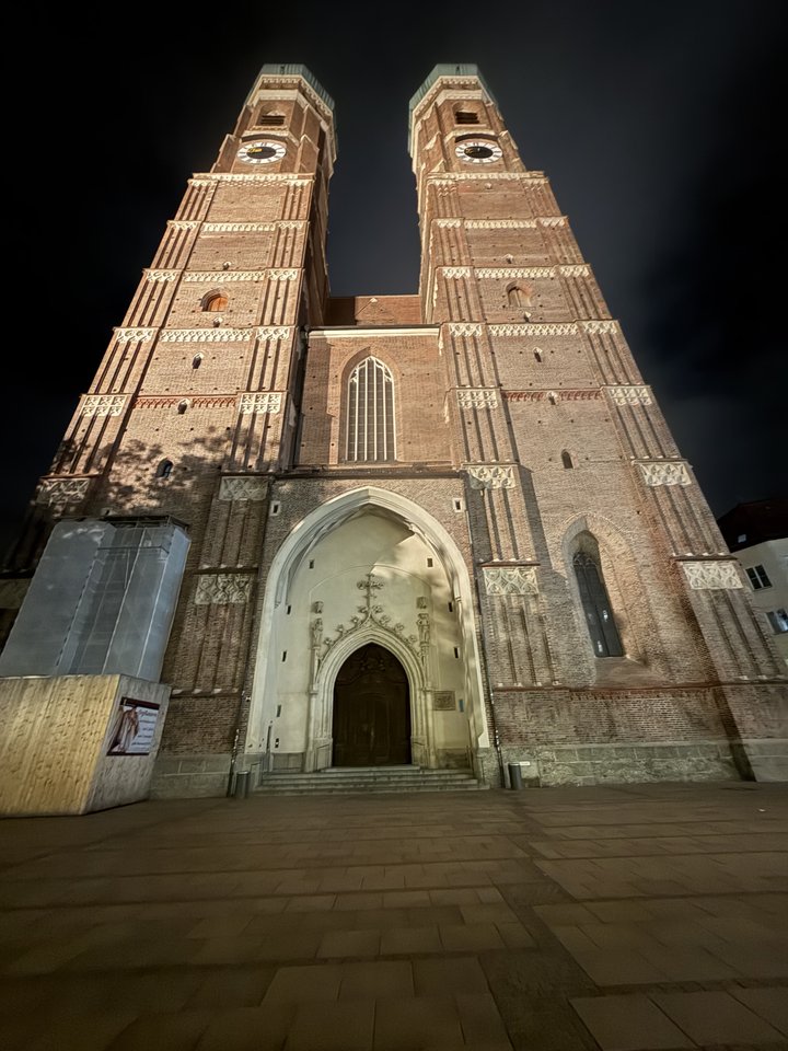 Low-angle night view of a tall Gothic brick cathedral with two towers and clock faces, lit warmly against a dark sky, with stone steps leading to an arched wooden doorway.