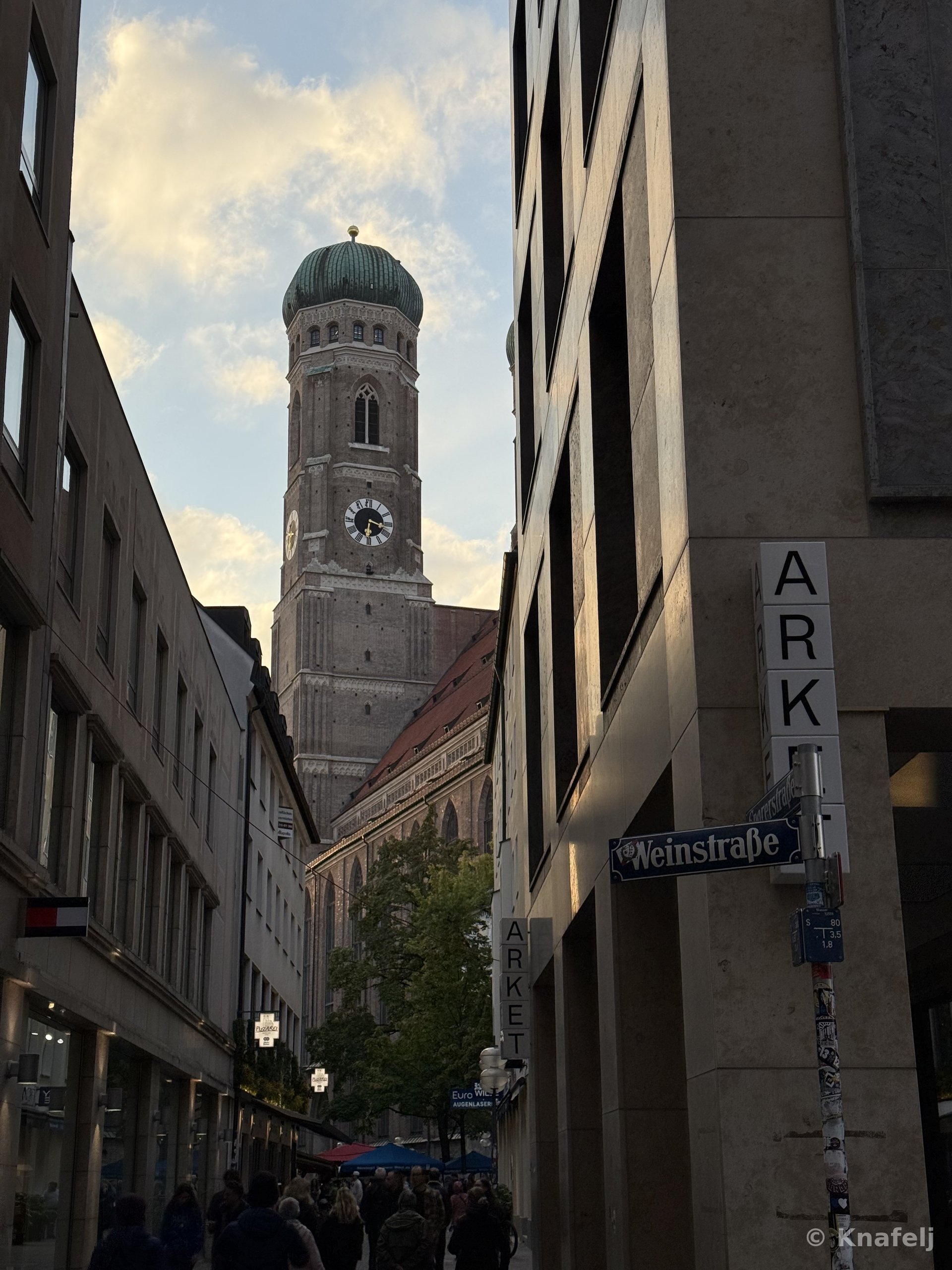 View down a narrow city street in Munich toward the brick tower and green dome of the Frauenkirche church, framed by buildings with street signs reading Weinstraße and Arket, with people walking below.