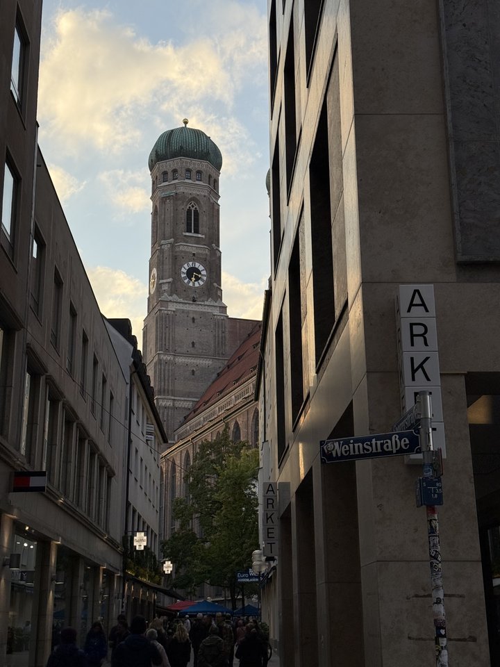 View down a narrow city street in Munich toward the brick tower and green dome of the Frauenkirche church, framed by buildings with street signs reading Weinstraße and Arket, with people walking below.