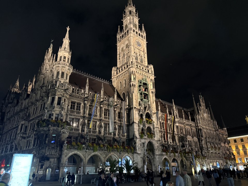 A grand Gothic town hall building lit up at night, featuring a tall central clock tower and ornate spires, with people walking in the plaza in the foreground and signage reading Marienplatz.
