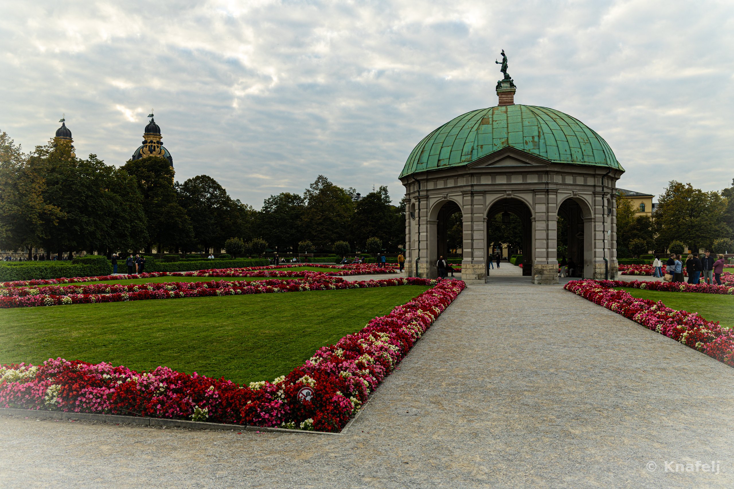 A stone pavilion with a green copper dome and a statue on top, surrounded by manicured lawns and geometric flower beds of red and pink blooms, with a gravel path leading to it and trees and domed towers in the background under a cloudy sky.