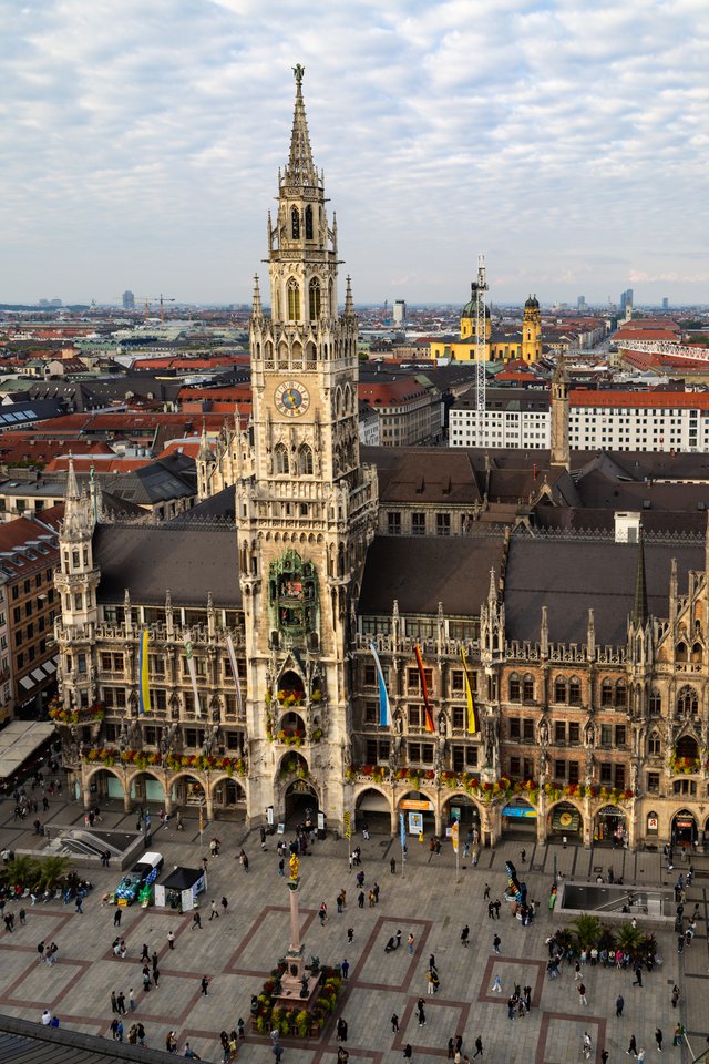 Aerial view of the New Town Hall in Munich with its tall Gothic spire and clock tower, people walking across the cobblestone square below, and red rooftops of the city extending under a cloudy sky.
