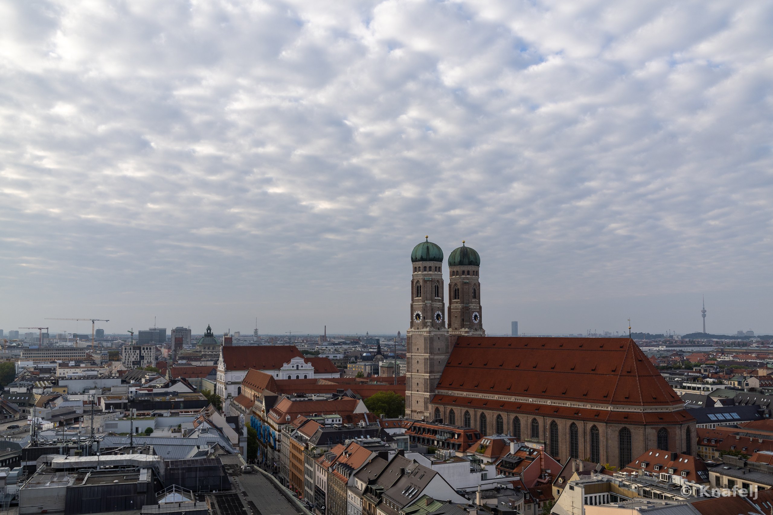 Aerial view of Munich showing the Frauenkirche cathedral with its two domed towers and large red roof, surrounded by rooftops of the city under a heavily clouded sky.