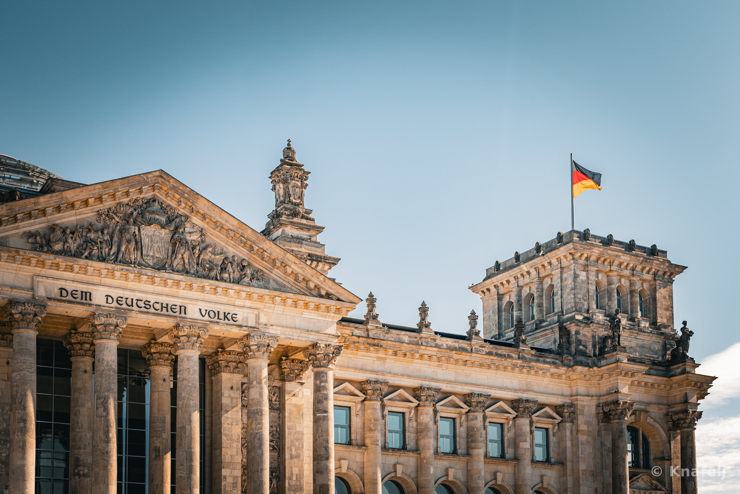 The Reichstag building in Berlin showing its columned portico with the inscription 'Dem Deutschen Volke', ornate stone carvings, and a German flag atop the corner tower.