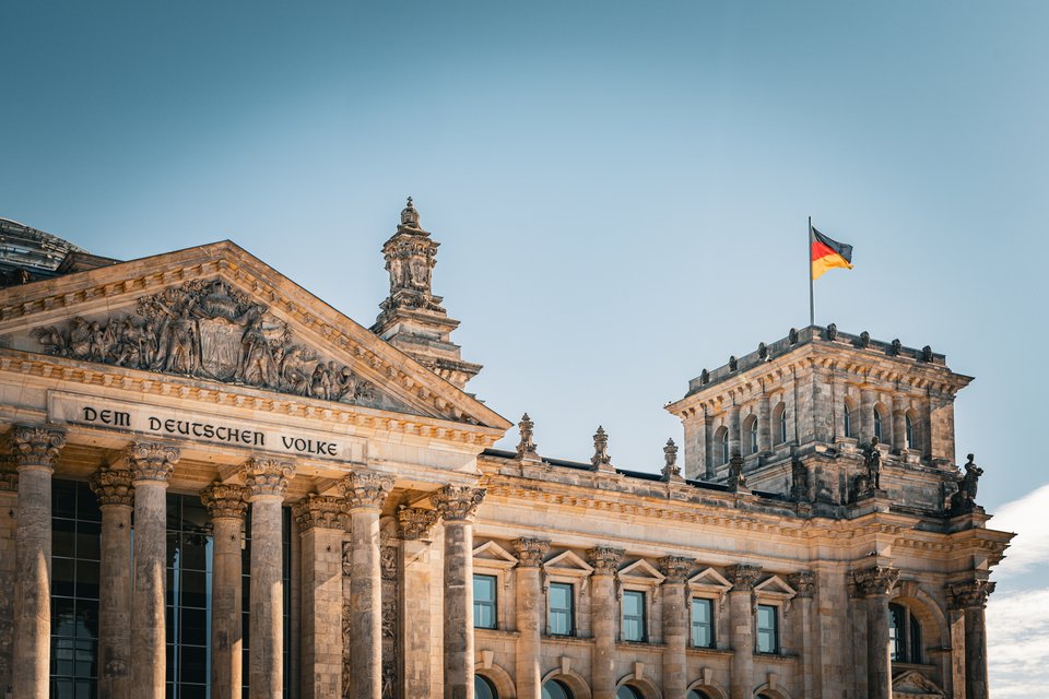 The Reichstag building in Berlin showing its columned portico with the inscription 'Dem Deutschen Volke', ornate stone carvings, and a German flag atop the corner tower.