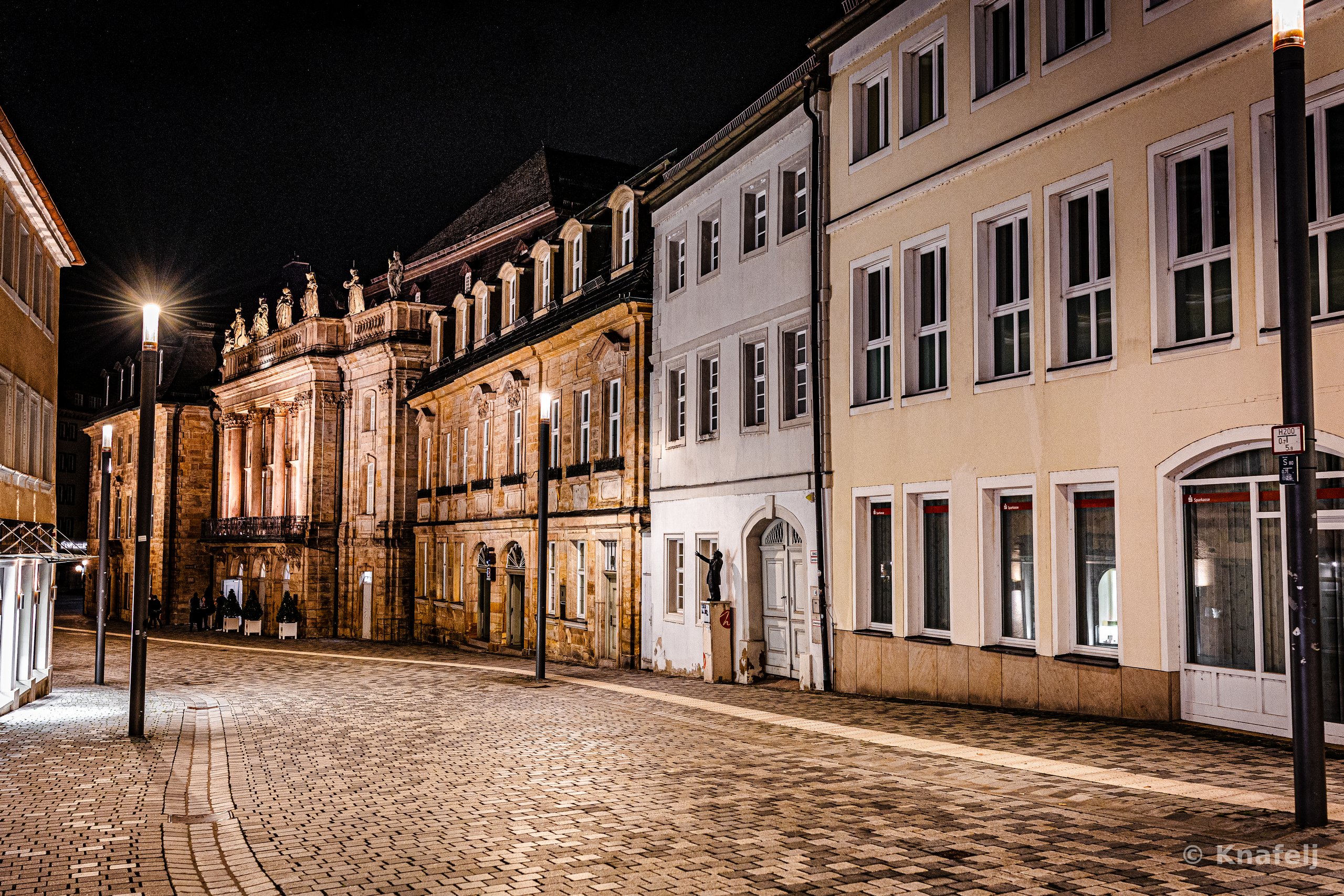 A deserted cobblestone street at night lined with historic European buildings, including an ornate baroque facade lit with warm light and tall street lamps glowing against a black sky.