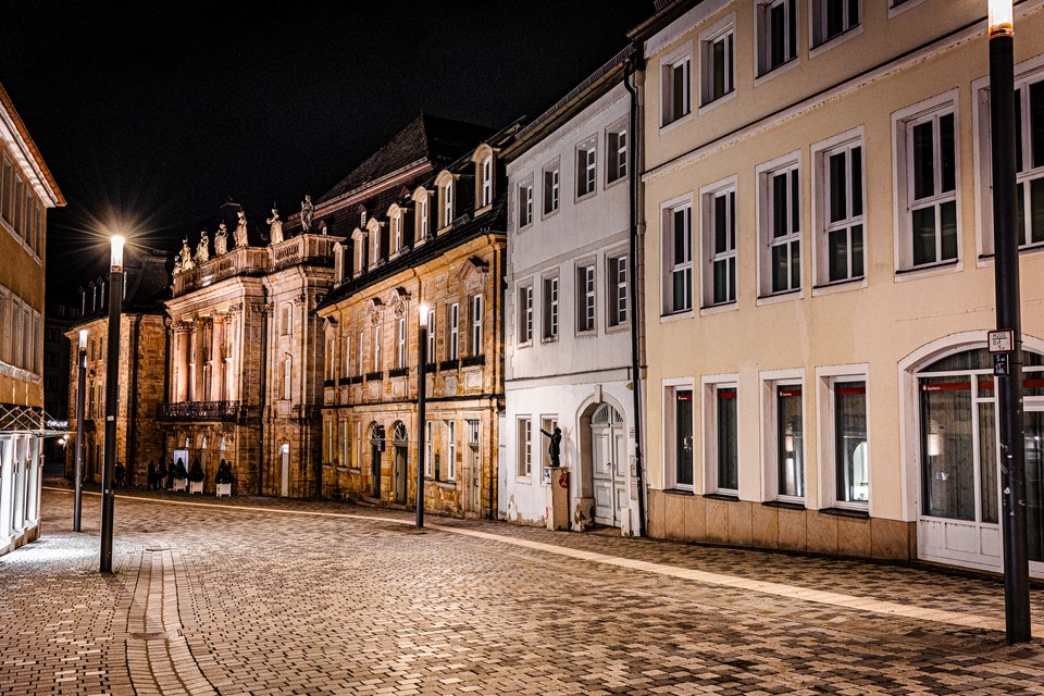 A deserted cobblestone street at night lined with historic European buildings, including an ornate baroque facade lit with warm light and tall street lamps glowing against a black sky.