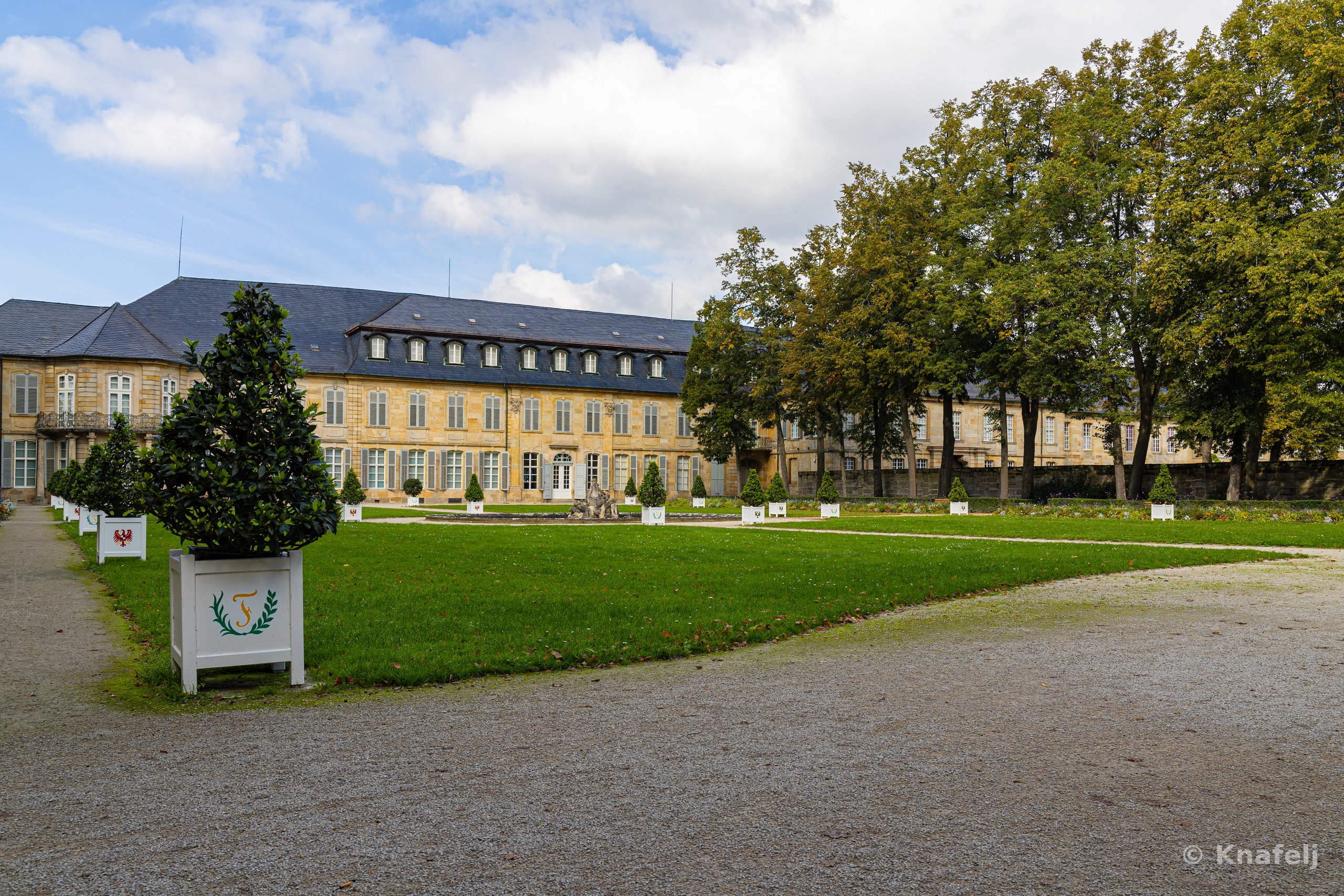 A large yellow baroque palace with slate roofs sits behind a green lawn and gravel courtyard, lined with white planter boxes holding cone-shaped topiary trees, surrounded by tall trees under a cloudy blue sky.