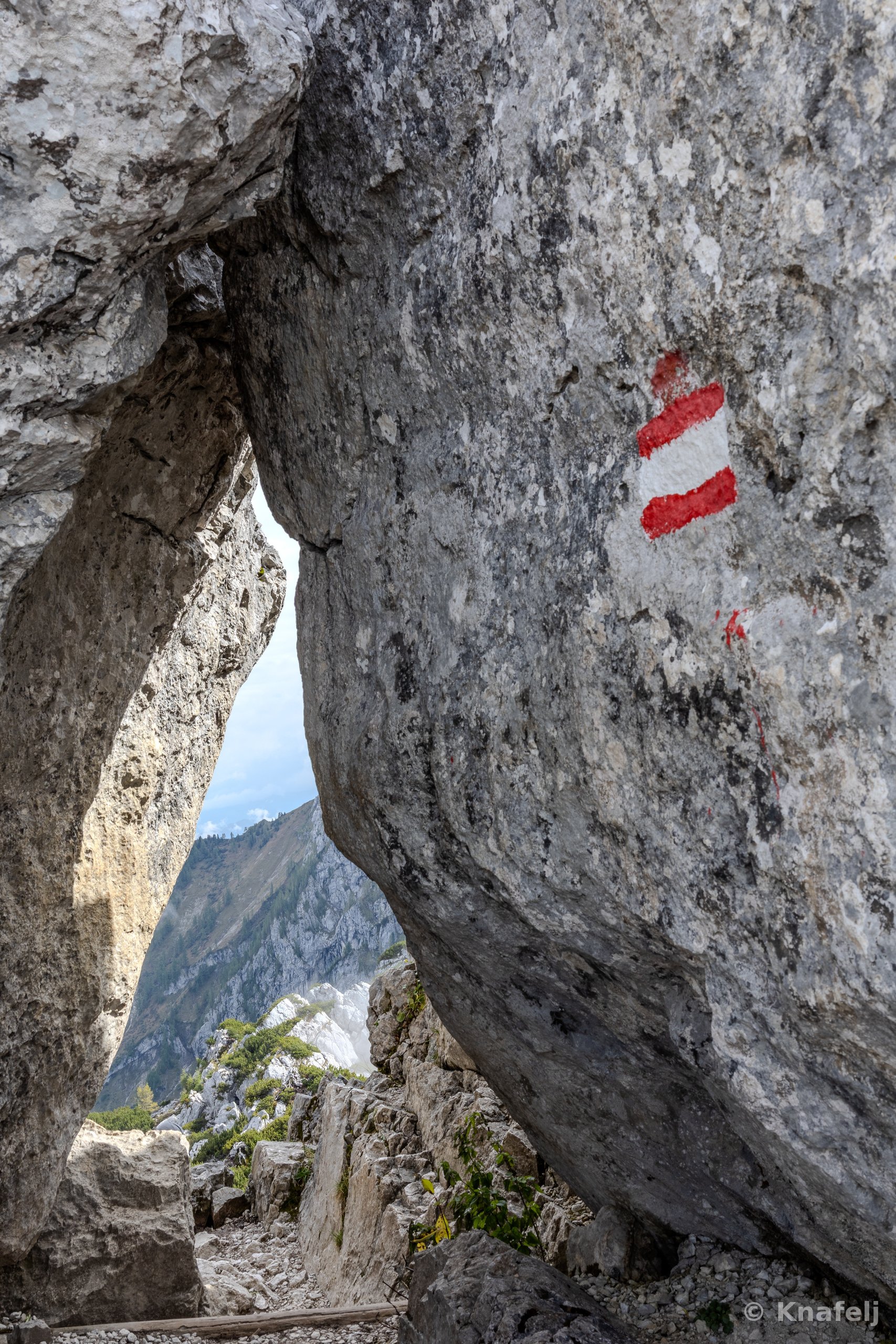 A large gray limestone rock face with a red-white-red painted hiking trail marker, beside a natural opening revealing distant mountain peaks and a rugged trail under a pale sky.