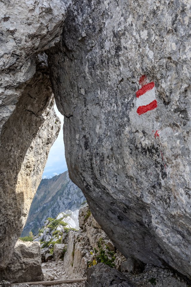 A large gray limestone rock face with a red-white-red painted hiking trail marker, beside a natural opening revealing distant mountain peaks and a rugged trail under a pale sky.