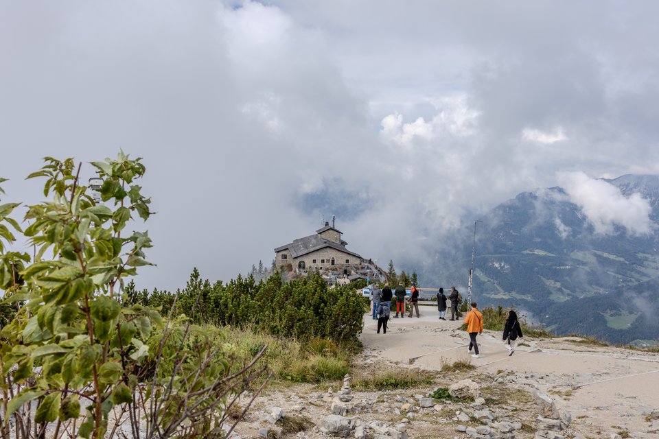 A stone building perched on a mountain ridge surrounded by clouds, with several people walking along a paved path lined by green shrubs and distant mountains in the background.