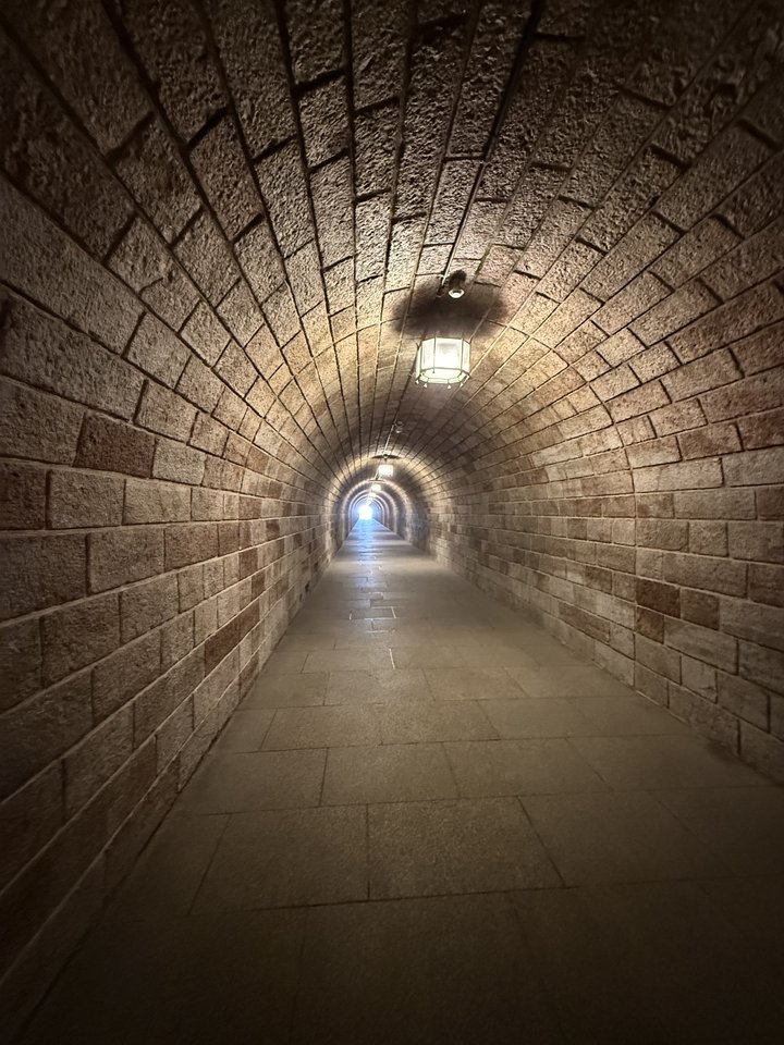 An arched underground tunnel built from stone bricks, with a tiled floor receding into the distance toward a small bright opening. A lantern hangs from the curved ceiling near the foreground.