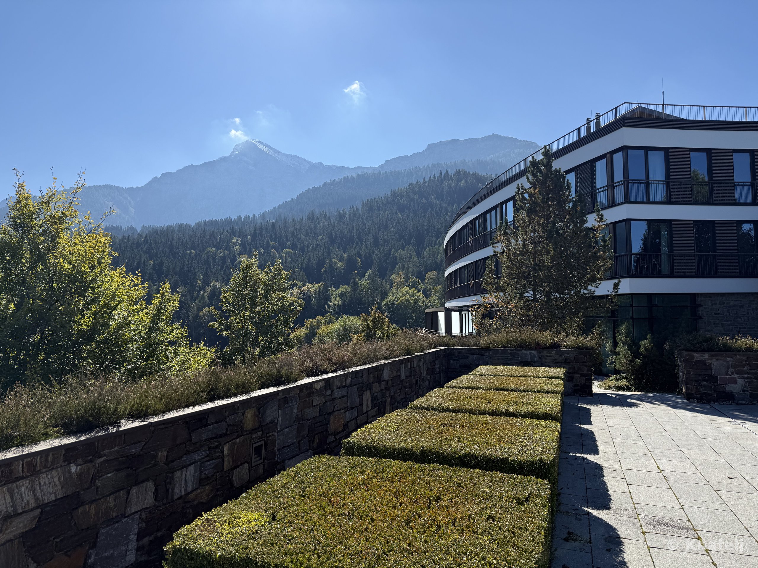 A contemporary multi-story building with balconies stands on the right, beside a terrace with rectangular green hedges bordered by a stone wall, overlooking a forested mountain range under a clear sky.