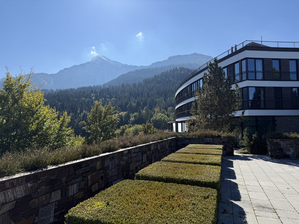 A contemporary multi-story building with balconies stands on the right, beside a terrace with rectangular green hedges bordered by a stone wall, overlooking a forested mountain range under a clear sky.