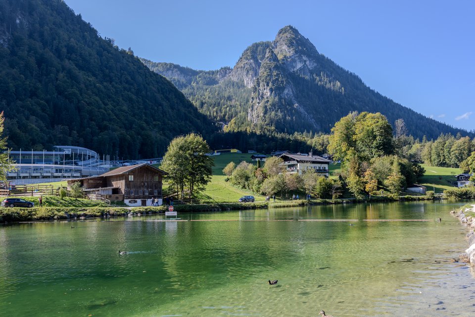 A clear green alpine lake with ducks swimming, backed by grassy meadows, wooden alpine buildings, dense forest, and a tall rocky mountain peak under a blue sky.