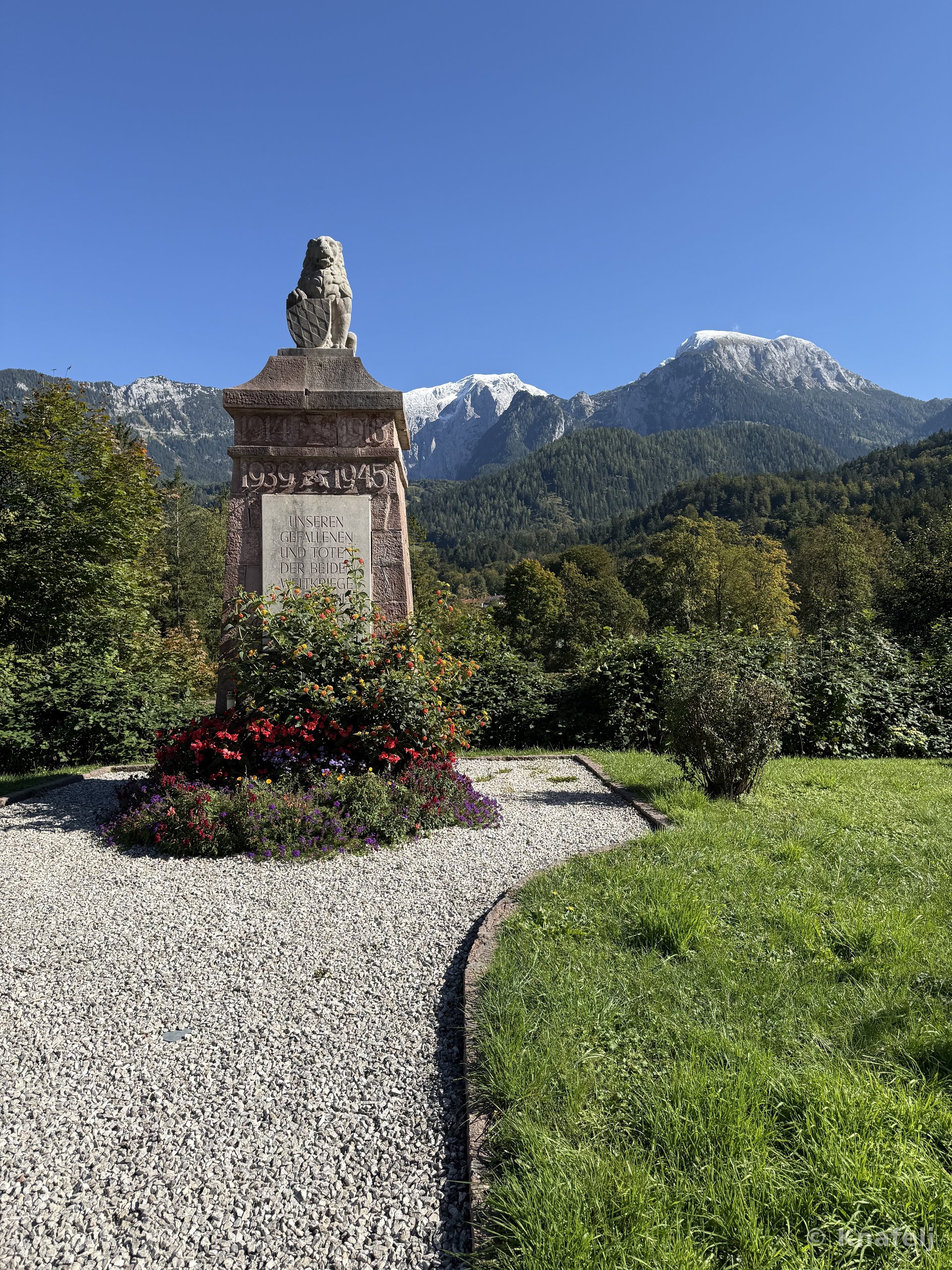 A stone war memorial topped with a carved lion, inscribed with dates 1939-1945, surrounded by red and purple flowers and a gravel path, with green forested mountains and snow-capped peaks in the background under a clear sky.