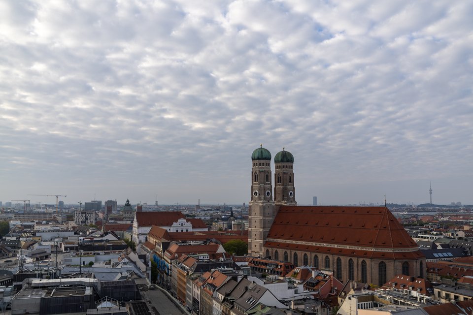 Aerial view of Munich showing the Frauenkirche cathedral with its two domed towers and large red roof, surrounded by rooftops of the city under a heavily clouded sky.