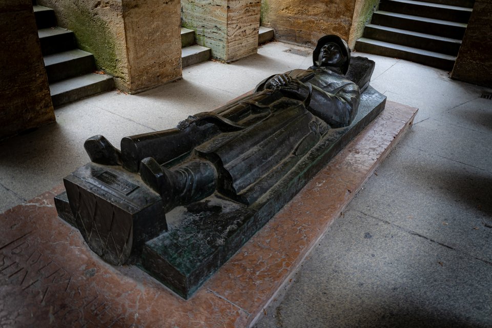 A dark bronze recumbent statue of a figure in robes resting on a marble tomb base, surrounded by stone steps and engraved floor inscriptions in a shadowy indoor space.