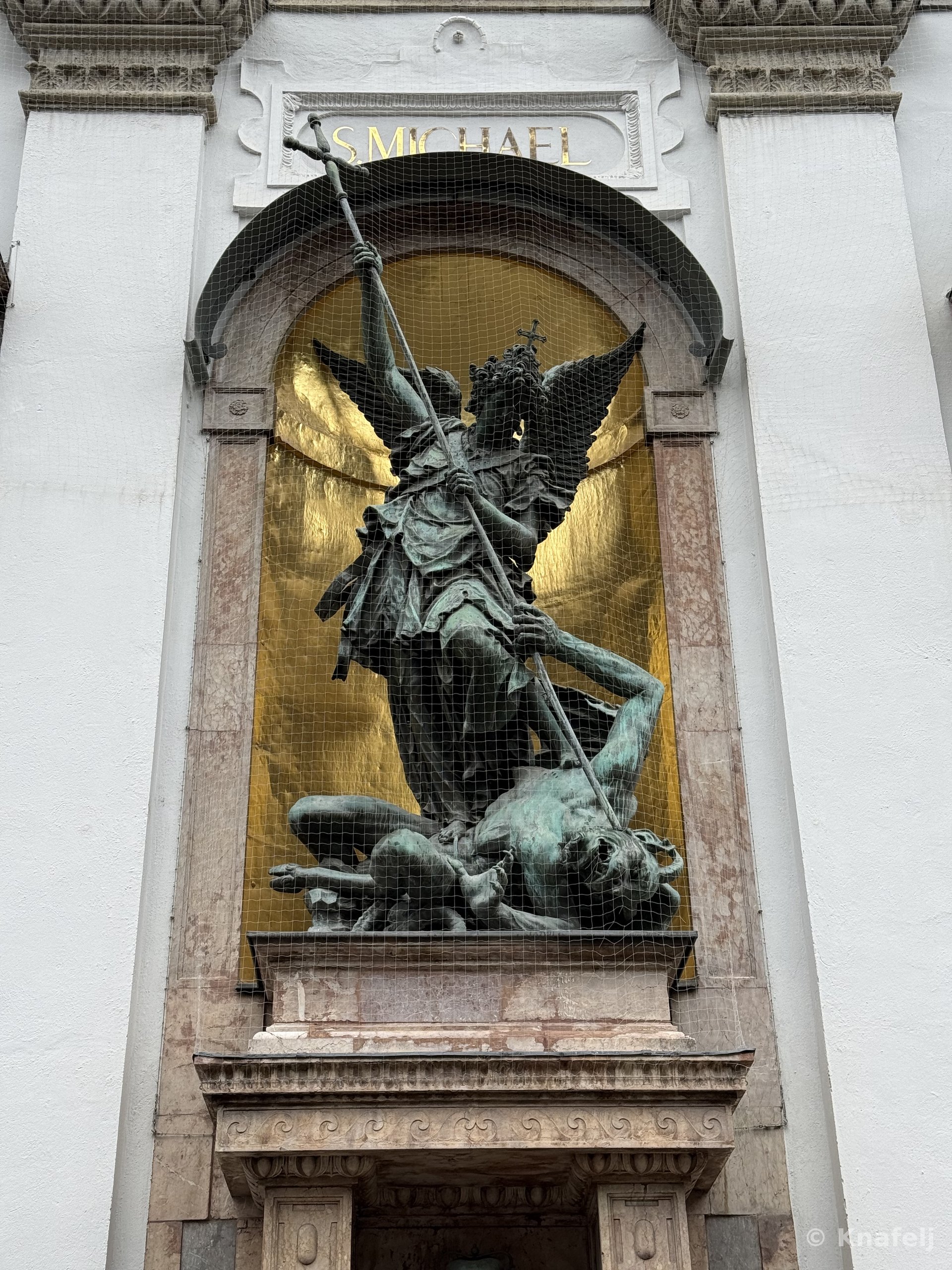 Bronze statue of the winged Archangel Michael in armor, holding a spear and standing over a fallen demon, displayed in an arched niche with a gold background and the inscription 'S. MICHAEL' above, on a white building facade.