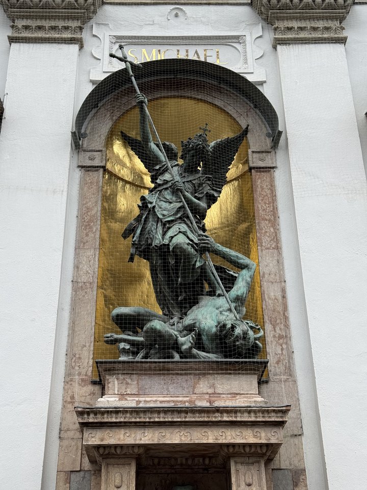 Bronze statue of the winged Archangel Michael in armor, holding a spear and standing over a fallen demon, displayed in an arched niche with a gold background and the inscription 'S. MICHAEL' above, on a white building facade.
