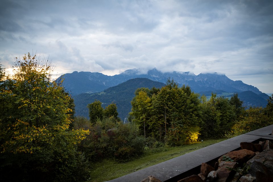 A mountain range partially hidden by low clouds at twilight, with dense green and yellowing trees in the foreground, a grassy lawn, and a stone wall in the lower right.