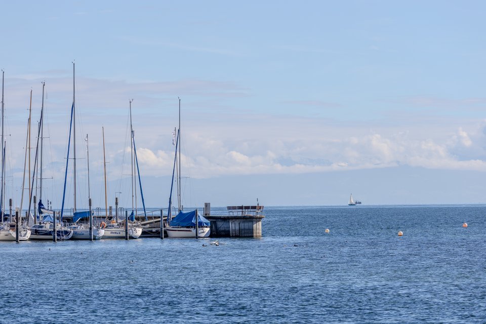 A marina with several moored sailboats and tall masts on the left, a small concrete dock structure, and calm blue water stretching to the horizon where a distant sailboat is visible under a partly cloudy sky.