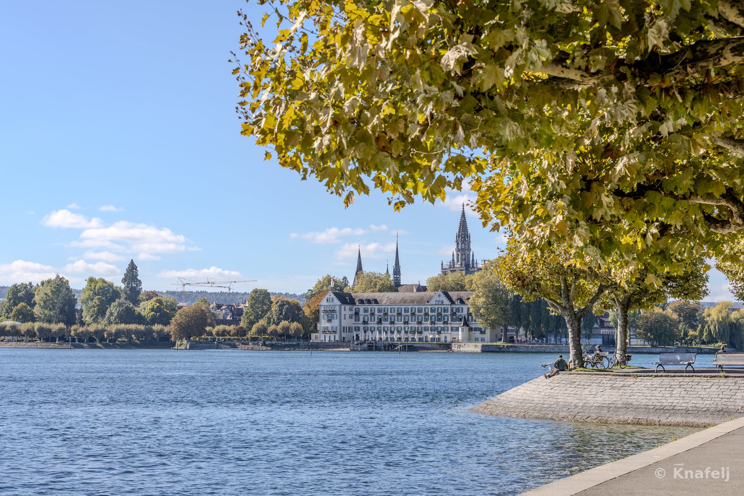 A lakeside promenade in autumn with trees bearing yellow-green leaves, looking across blue water toward a historic town skyline featuring church spires and a large white building, under a blue sky with scattered clouds.