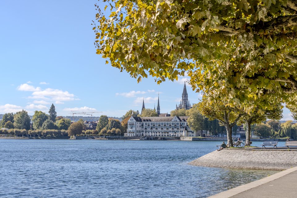 A lakeside promenade in autumn with trees bearing yellow-green leaves, looking across blue water toward a historic town skyline featuring church spires and a large white building, under a blue sky with scattered clouds.