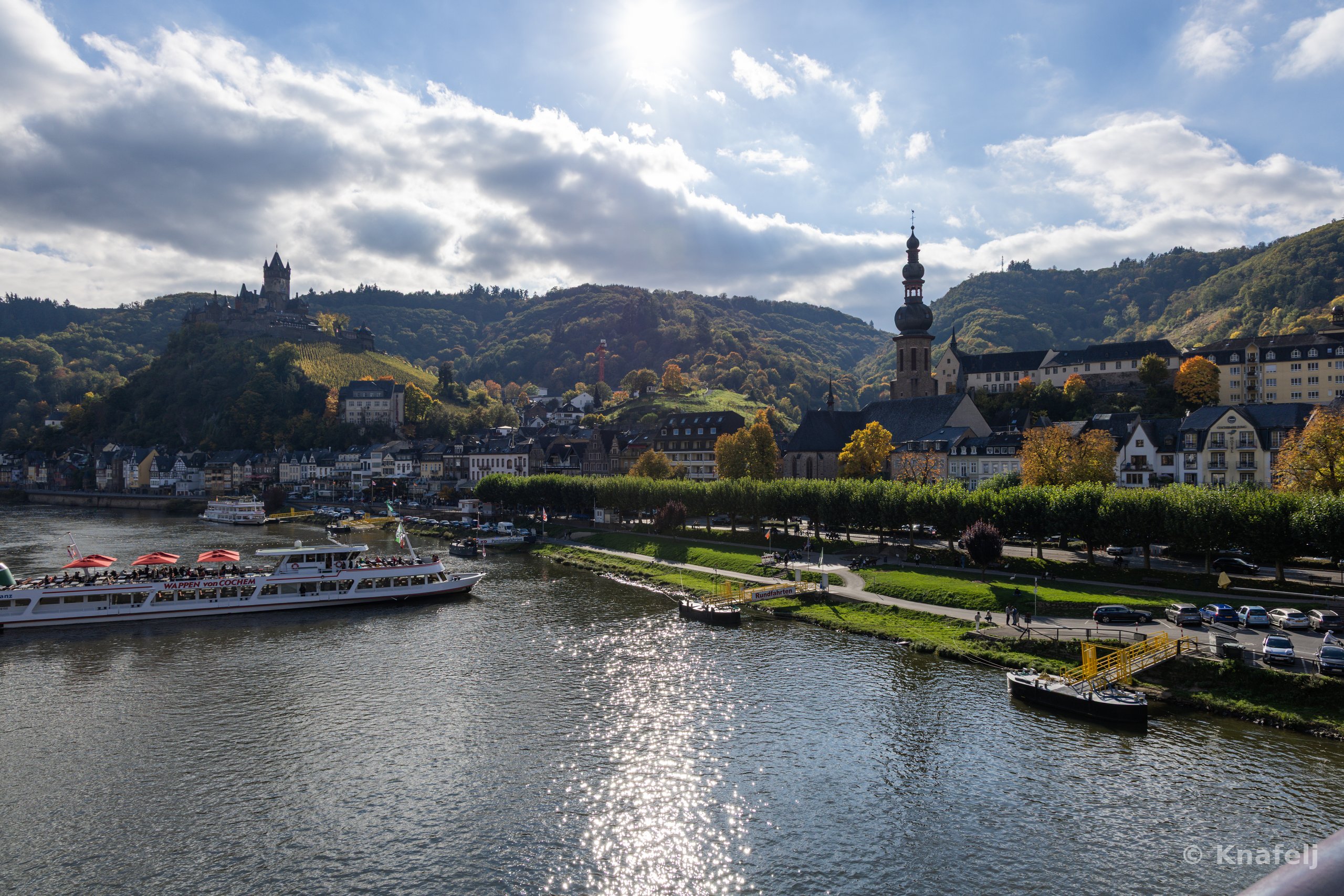 A wide view of a German riverside town along the Moselle River, with a white tourist boat on the left, a church spire and town buildings in the center, a hilltop castle on the left ridge, and forested hills under a bright cloudy sky.