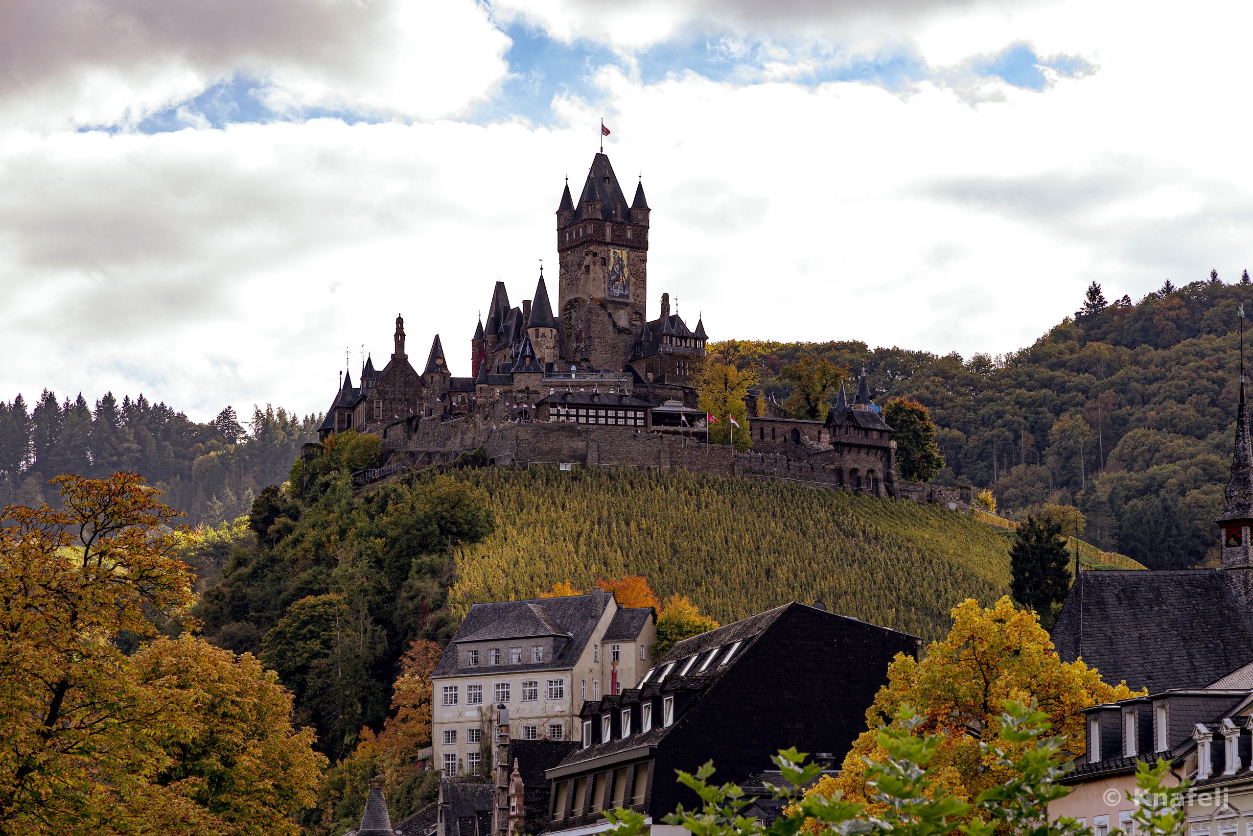 A medieval German castle with tall spires and a clock tower sits atop a green hill covered with trees turning golden in autumn, with town buildings below and a cloudy sky overhead.