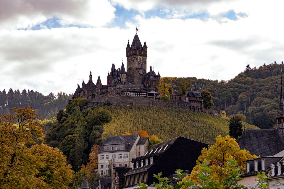 A medieval German castle with tall spires and a clock tower sits atop a green hill covered with trees turning golden in autumn, with town buildings below and a cloudy sky overhead.