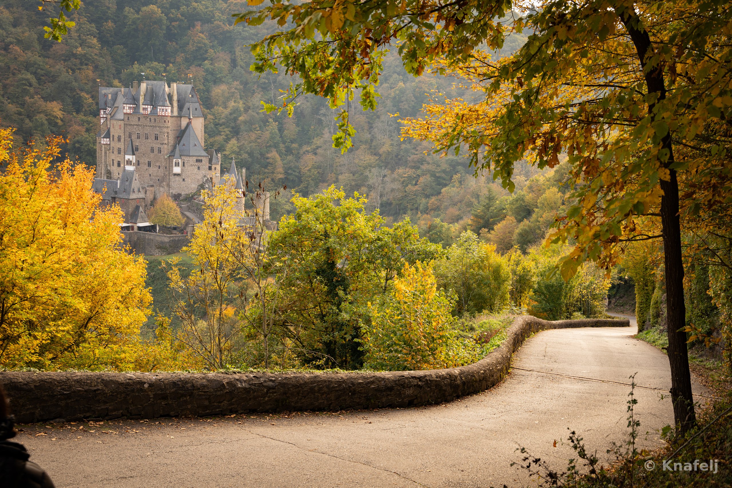A fairytale-like stone castle on a hill surrounded by trees with green and yellow autumn foliage, while a paved road bordered by a low stone wall winds through the foreground.