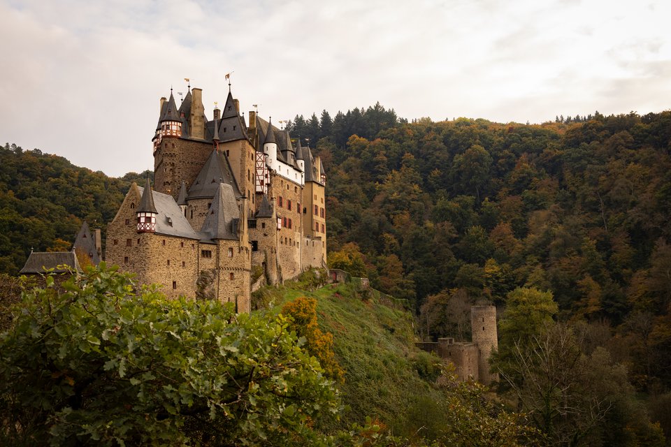 A large medieval German castle with multiple spires and stone towers set on a wooded hill, surrounded by green and autumn-colored forest under a cloudy sky.