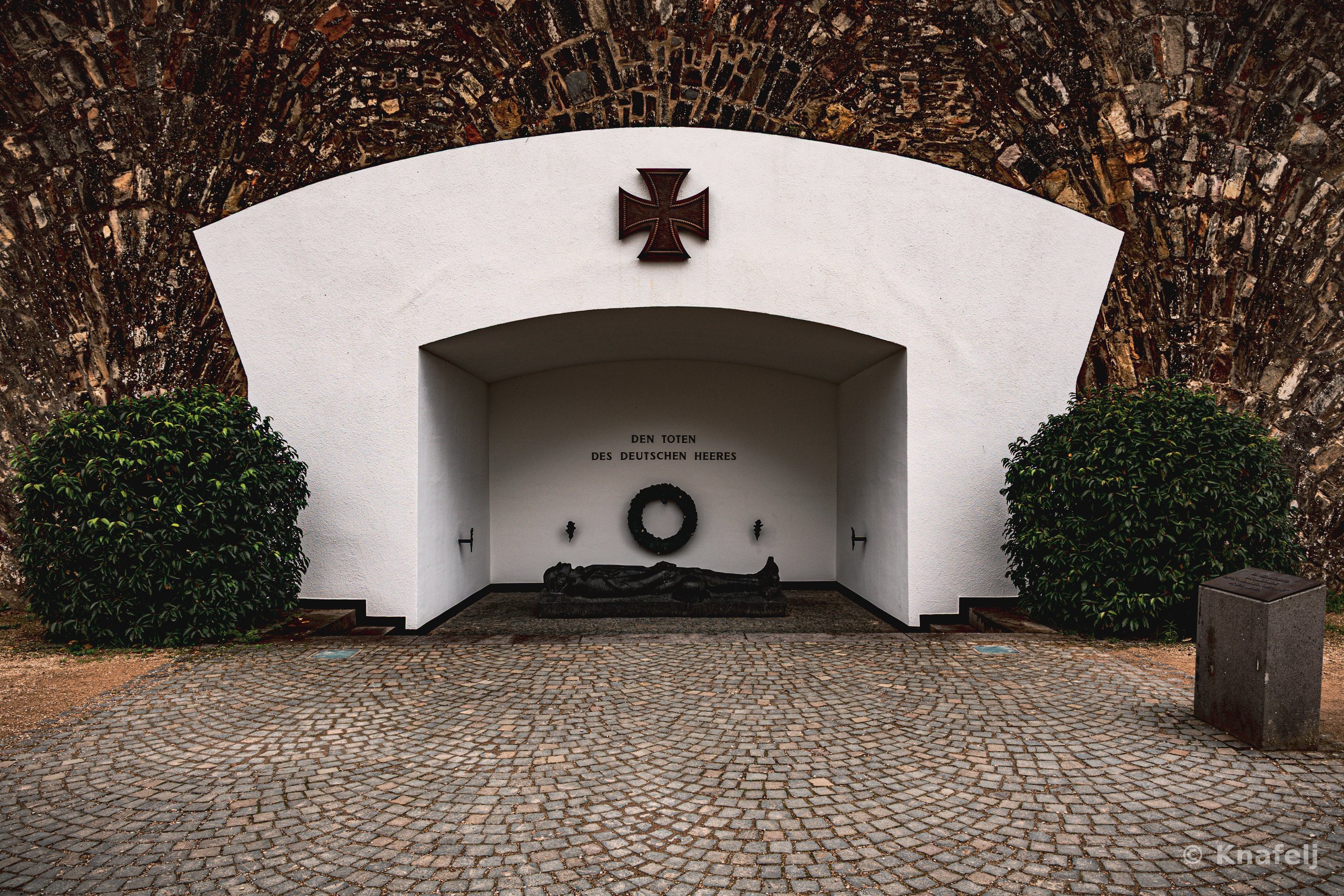 A white memorial structure built into a stone archway, displaying a dark Iron Cross above an inscription reading 'Den Toten des Deutschen Heeres,' with a wreath and a reclining sculpted figure inside, flanked by two rounded green bushes over a cobblestone courtyard.